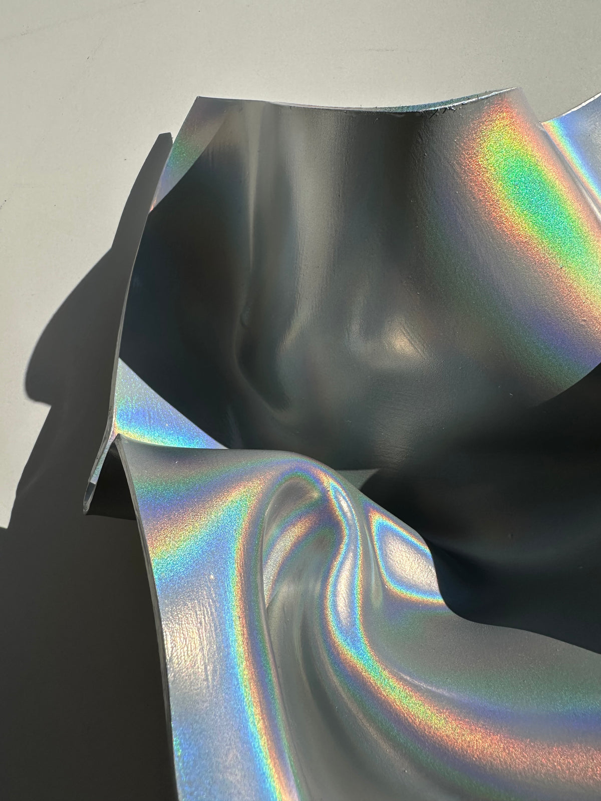 A close-up of a sheet of holographic silver vinyl or adhesive film, showing its reflective iridescent surface with rainbow colors shifting across a dark gray base.