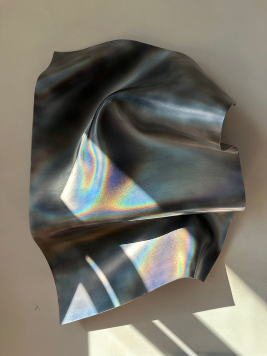 An abstract, crumpled sheet of iridescent metallic wall sculpture art with a holographic finish, casting colorful reflections under direct sunlight against a neutral background.