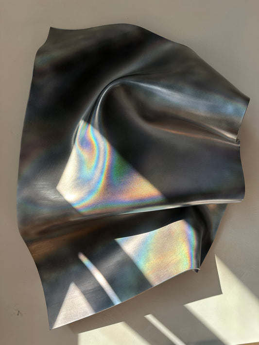 An iridescent holographic sculptural art draped in soft folds, showing a dark gray base with rainbow reflections under natural light.