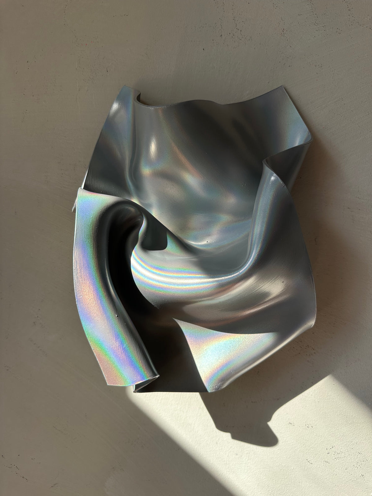 An abstract sculptural wall art piece made of metallic, crumpled material with an iridescent silver finish, mounted on a textured beige wall with dramatic sunlight casting shadows.