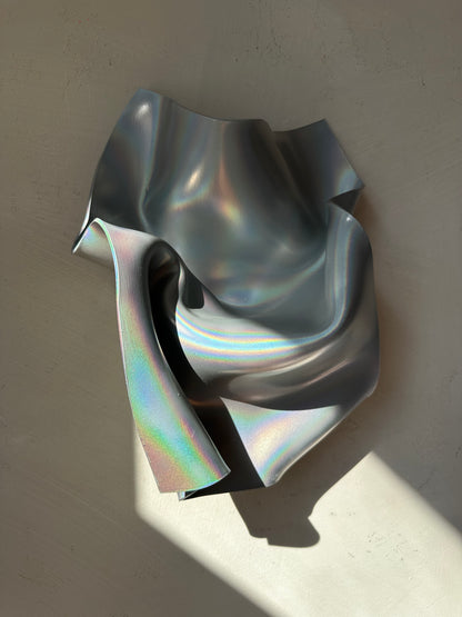 An abstract sculptural wall art piece made of iridescent, metallic silver material with flowing folds and a reflective finish, mounted against a textured beige wall with a strong diagonal shadow.