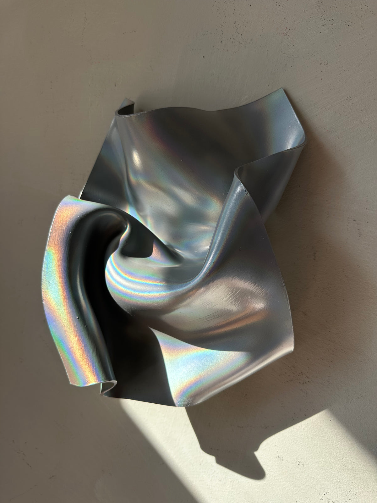 An abstract sculptural wall art piece made of iridescent metallic material, draped in flowing folds against a light beige wall with natural sunlight casting shadows.