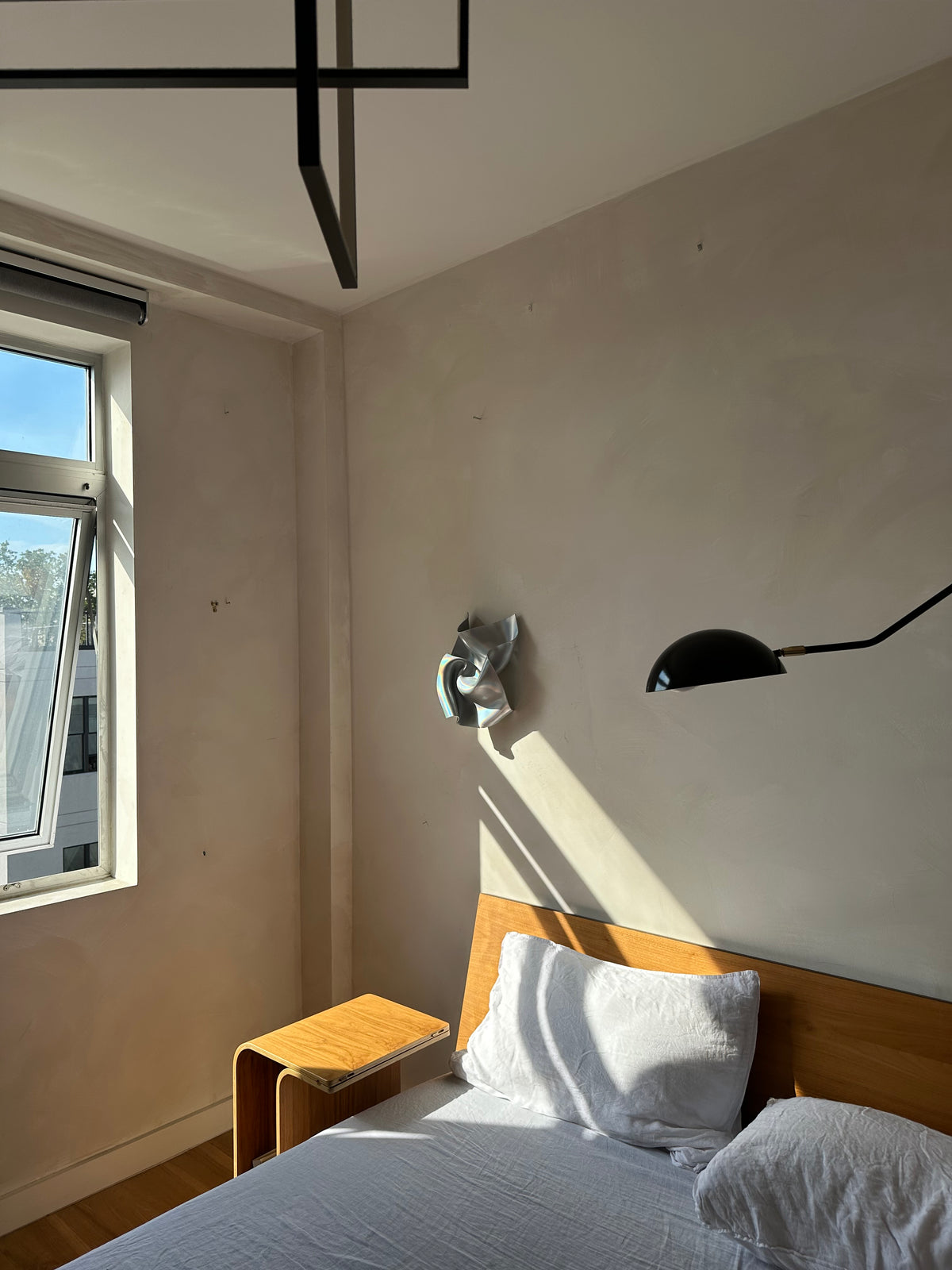 A modern bedroom corner with a bed, white pillows, a wooden headboard, a small wooden side table, a black wall lamp, and a window casting strong sunlight on the beige wall.