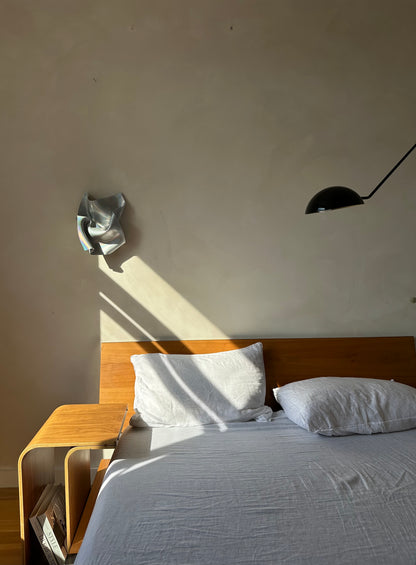 A minimalist bedroom scene featuring a bed with white linens and a wooden headboard, set against a textured beige wall with dramatic natural sunlight casting shadows. A black dome-shaped wall lamp is mounted on the wall, and a small wooden side table sits beside the bed.