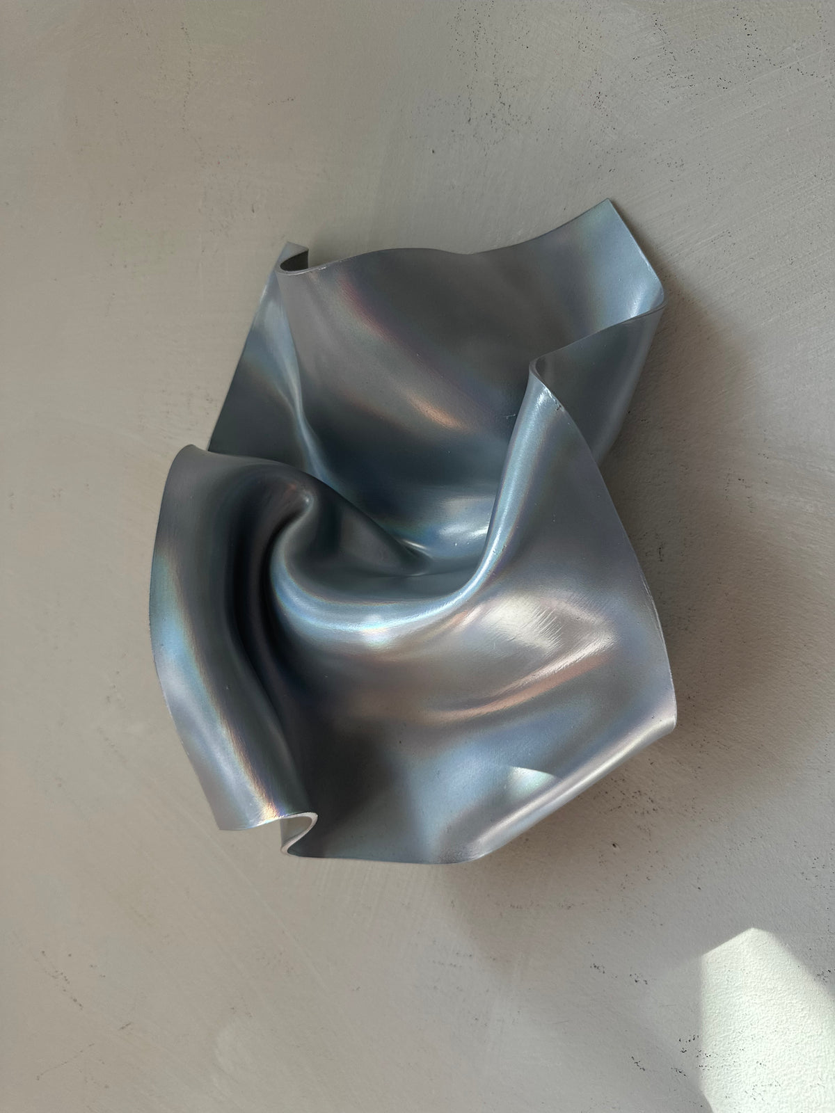 A sculptural, metallic silver wall-mounted piece with fluid, crumpled folds and a reflective finish, set against a textured light gray wall.