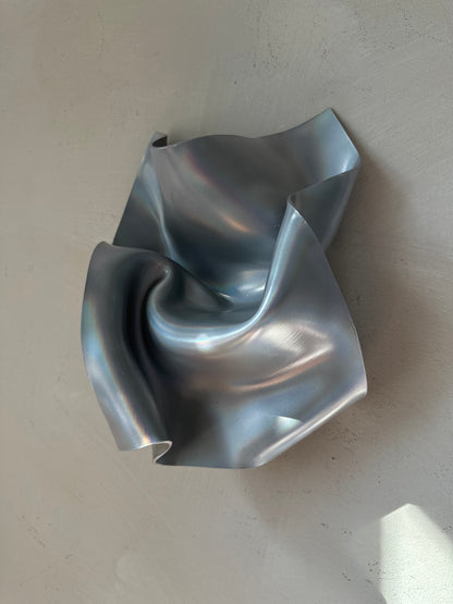 A sculptural, metallic silver wall-mounted piece with fluid, crumpled folds and a reflective finish, set against a textured light gray wall.