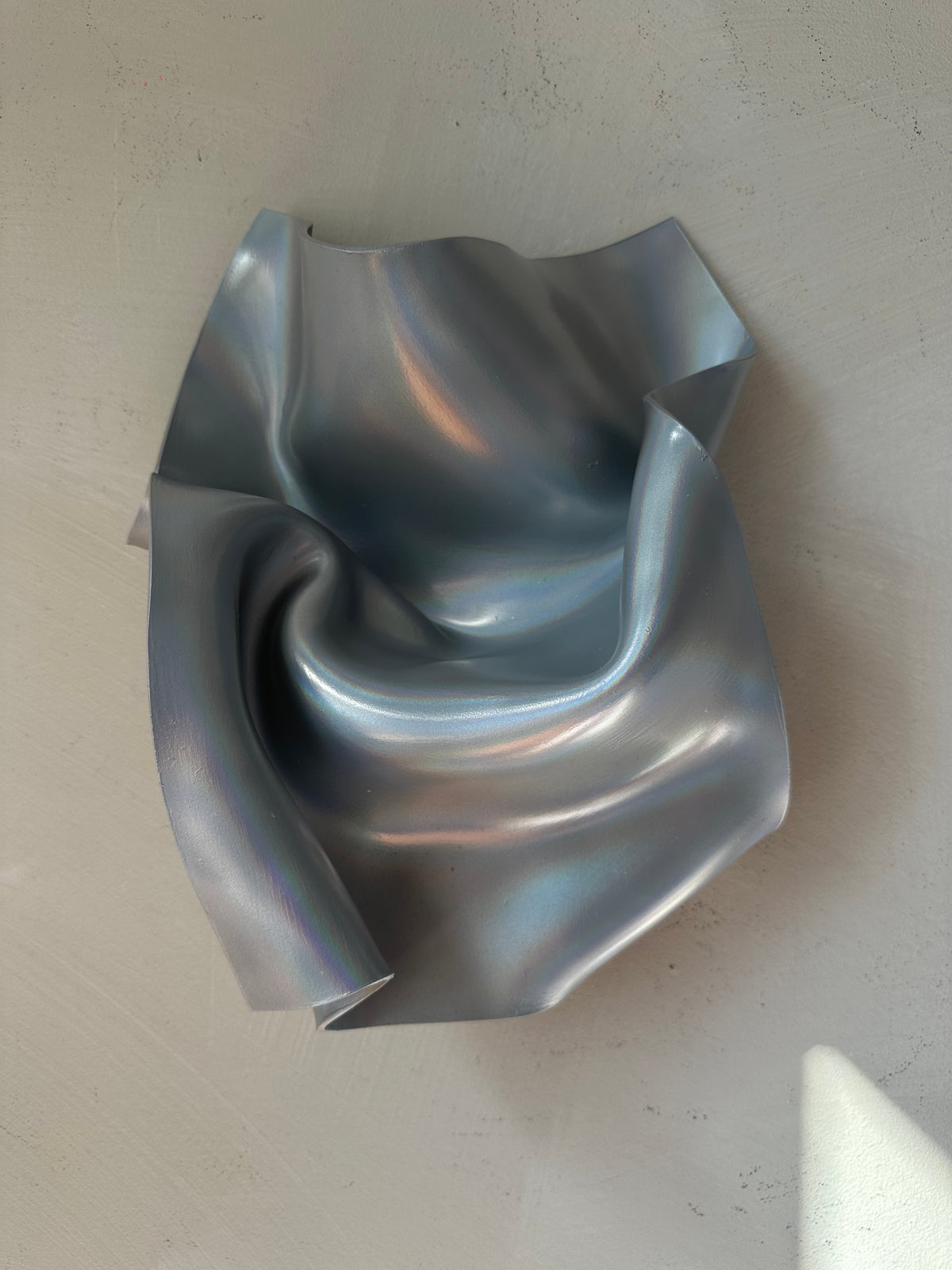 A sculptural, metallic silver bowl with a crumpled, organic texture, photographed from a top-down perspective on a neutral, light gray textured surface.