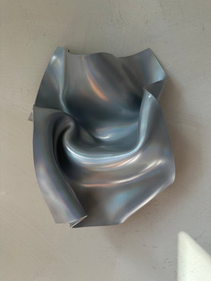 A sculptural, metallic silver bowl with a crumpled, organic texture, photographed from a top-down perspective on a neutral, light gray textured surface.