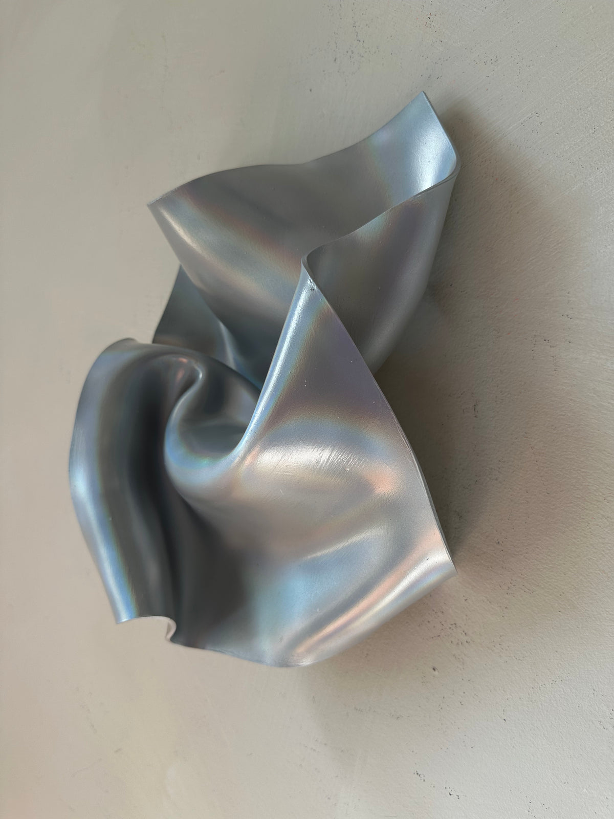 A sculptural metallic wall art piece with a crumpled, flowing form in a reflective silver finish, mounted against a plain off-white wall.
