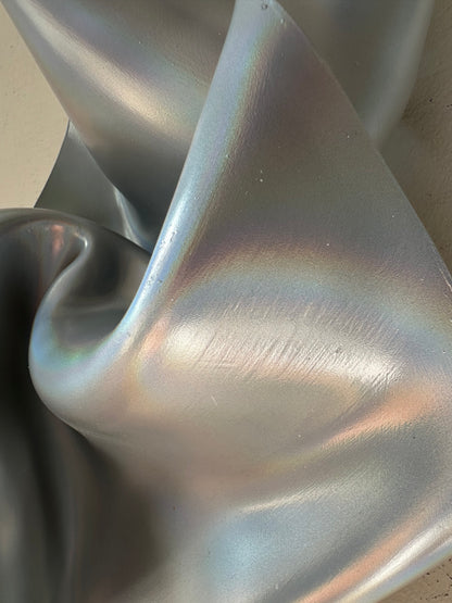 A close-up of draped holographic silver fabric with an iridescent finish reflecting pastel rainbow tones.