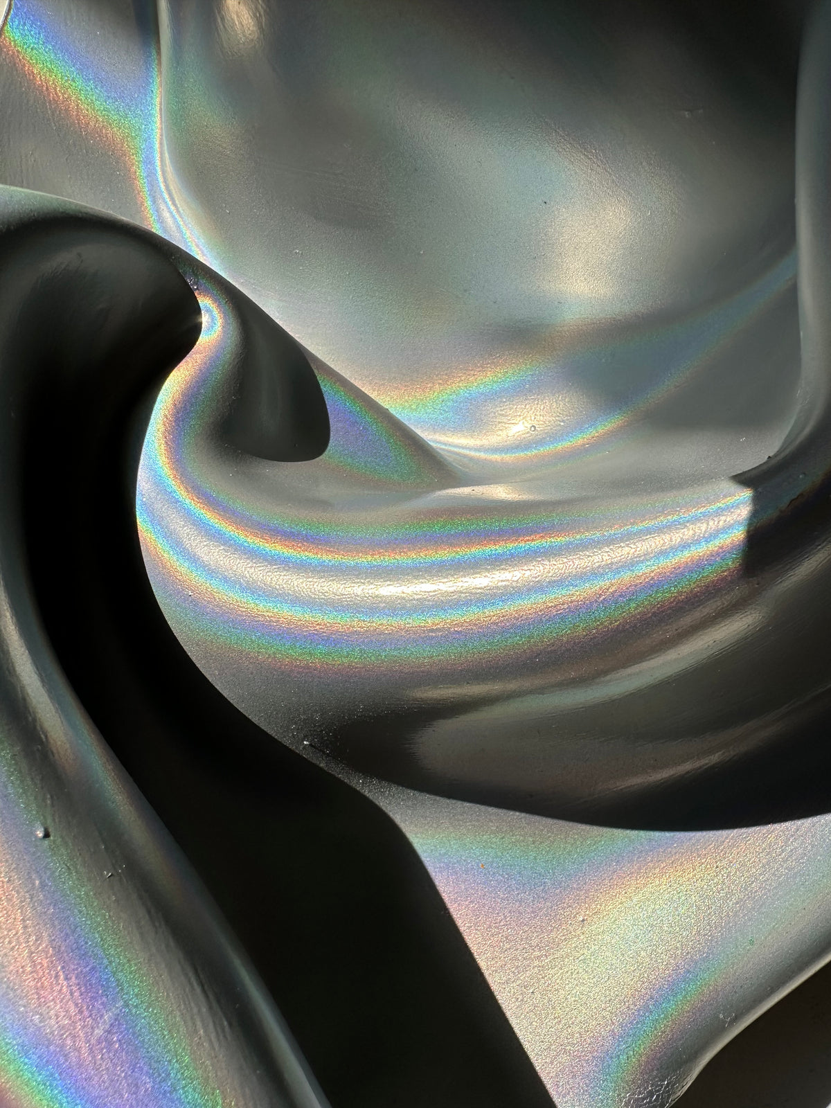 A close-up of a dark, iridescent, fluid-like surface with shifting rainbow reflections and deep black shadows.