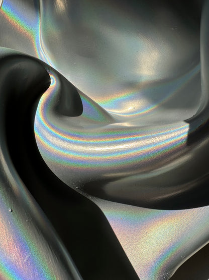 A close-up of a dark, iridescent, fluid-like surface with shifting rainbow reflections and deep black shadows.
