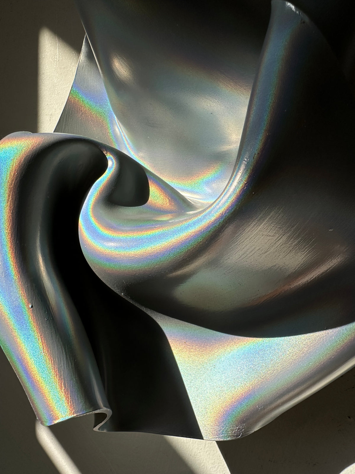 A close-up of a dark, metallic, iridescent material with flowing folds and rainbow reflections under directional light.